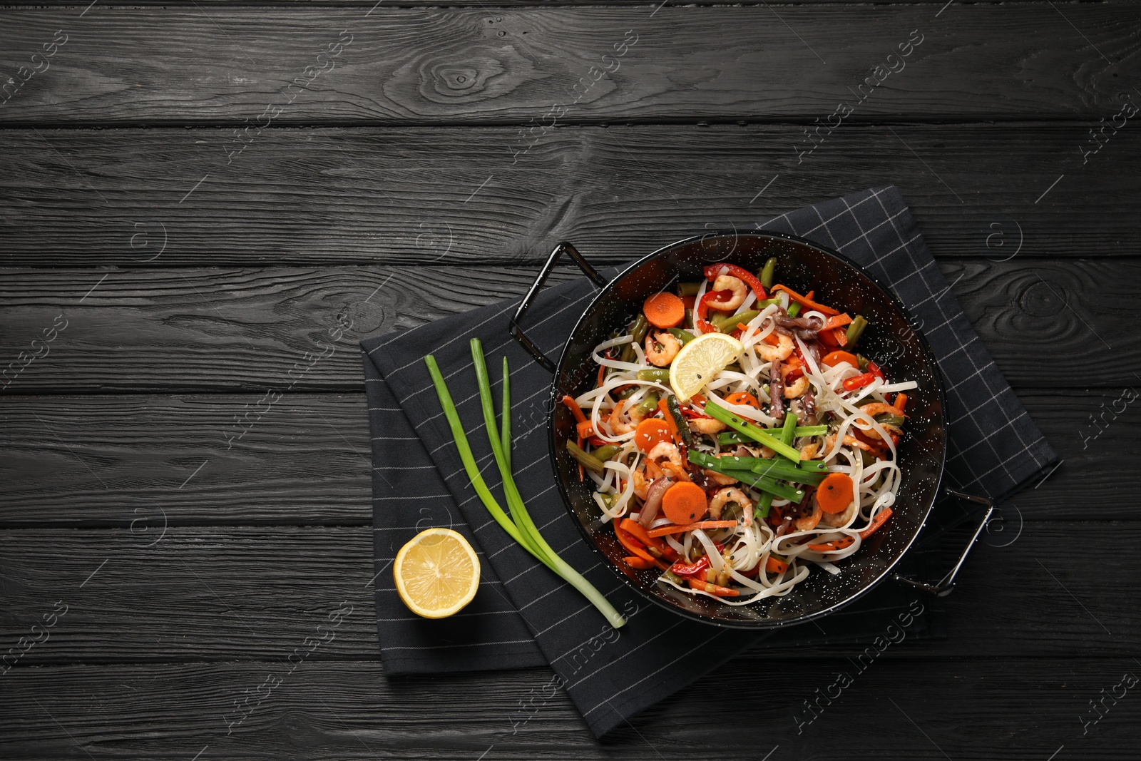 Photo of Shrimp stir fry with noodles and vegetables in wok on black wooden table, top view. Space for text