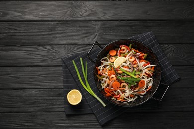 Photo of Shrimp stir fry with noodles and vegetables in wok on black wooden table, top view. Space for text