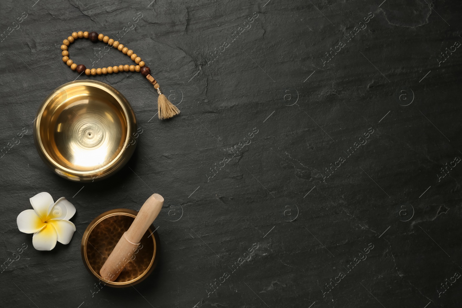 Photo of Golden singing bowls with mallet, beads and flower on black table, flat lay. Space for text