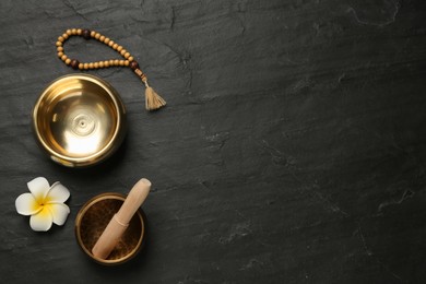Photo of Golden singing bowls with mallet, beads and flower on black table, flat lay. Space for text
