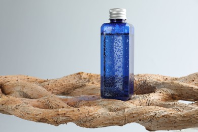 Bottle of cosmetic product on wooden branch against light grey background, space for text