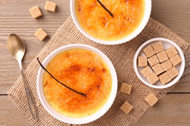 Photo of Delicious creme brulee in bowls, vanilla pods and sugar cubes on wooden table, top view