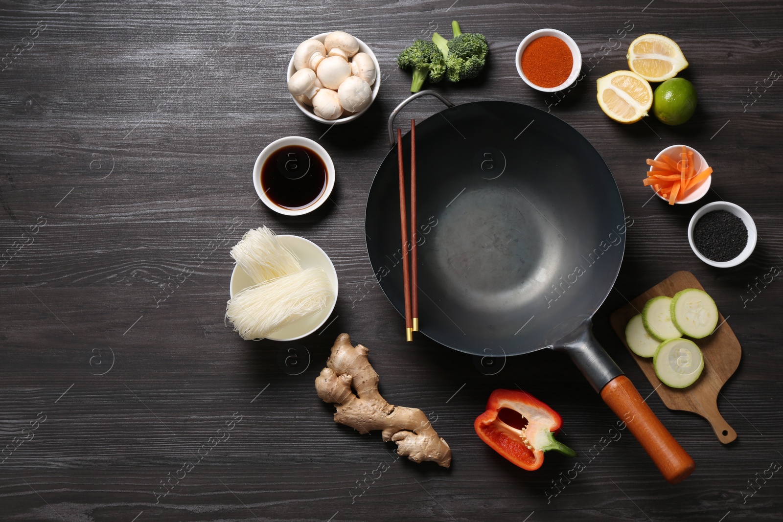 Photo of Empty iron wok and chopsticks surrounded by ingredients on dark grey wooden table, flat lay. Space for text