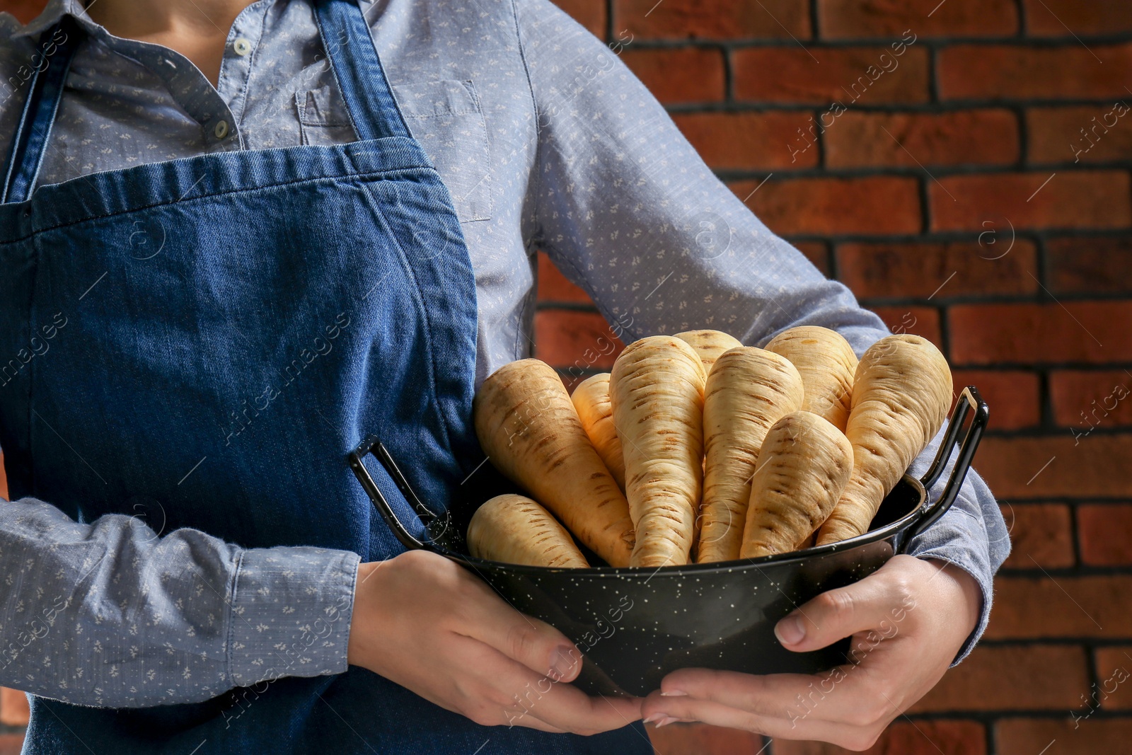 Photo of Woman holding bowl with fresh ripe parsnips near red brick wall, closeup