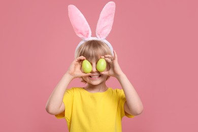 Photo of Happy boy in bunny ears headband holding painted Easter eggs near his eyes on pink background