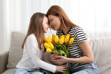 Mother and her cute daughter with bouquet of yellow tulips on sofa at home
