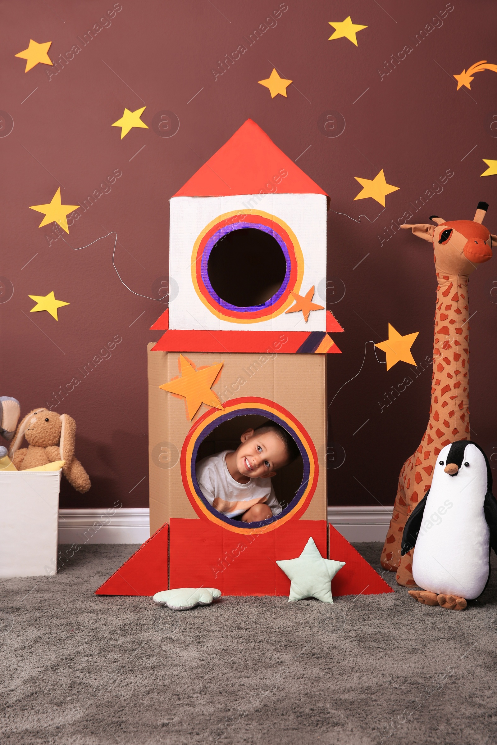 Photo of Cute little boy playing with cardboard rocket and toys at home. Child's room interior