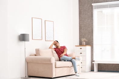 Photo of Young man with air conditioner remote control suffering from heat on sofa at home