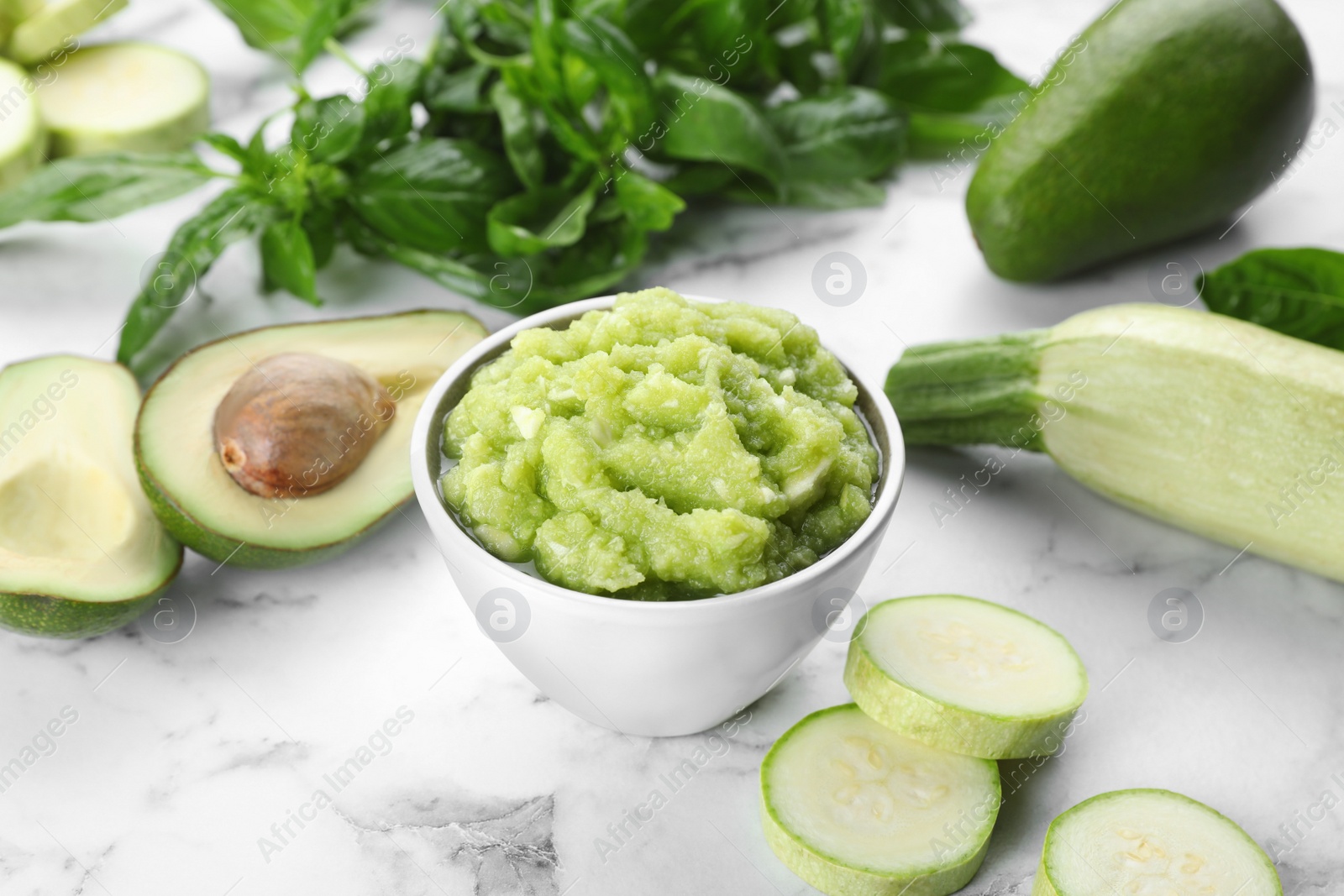 Photo of Bowl with tasty puree, zucchini, avocado and basil on white marble table