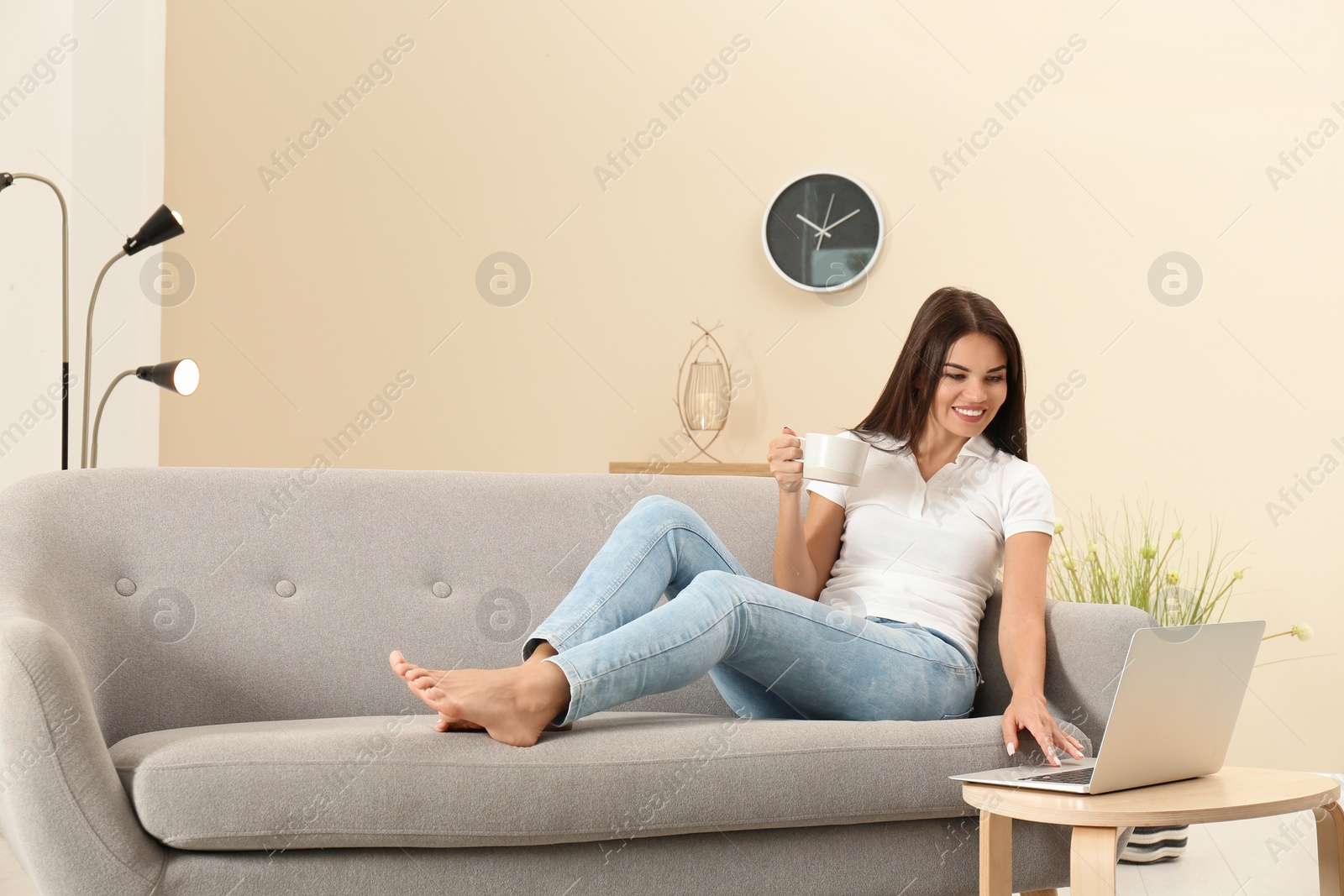 Photo of Beautiful woman with cup and laptop sitting on sofa at home