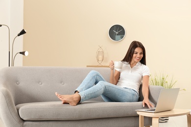 Beautiful woman with cup and laptop sitting on sofa at home