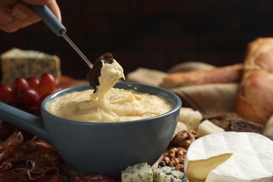 Photo of Woman dipping piece of bread into fondue pot with melted cheese at table, closeup