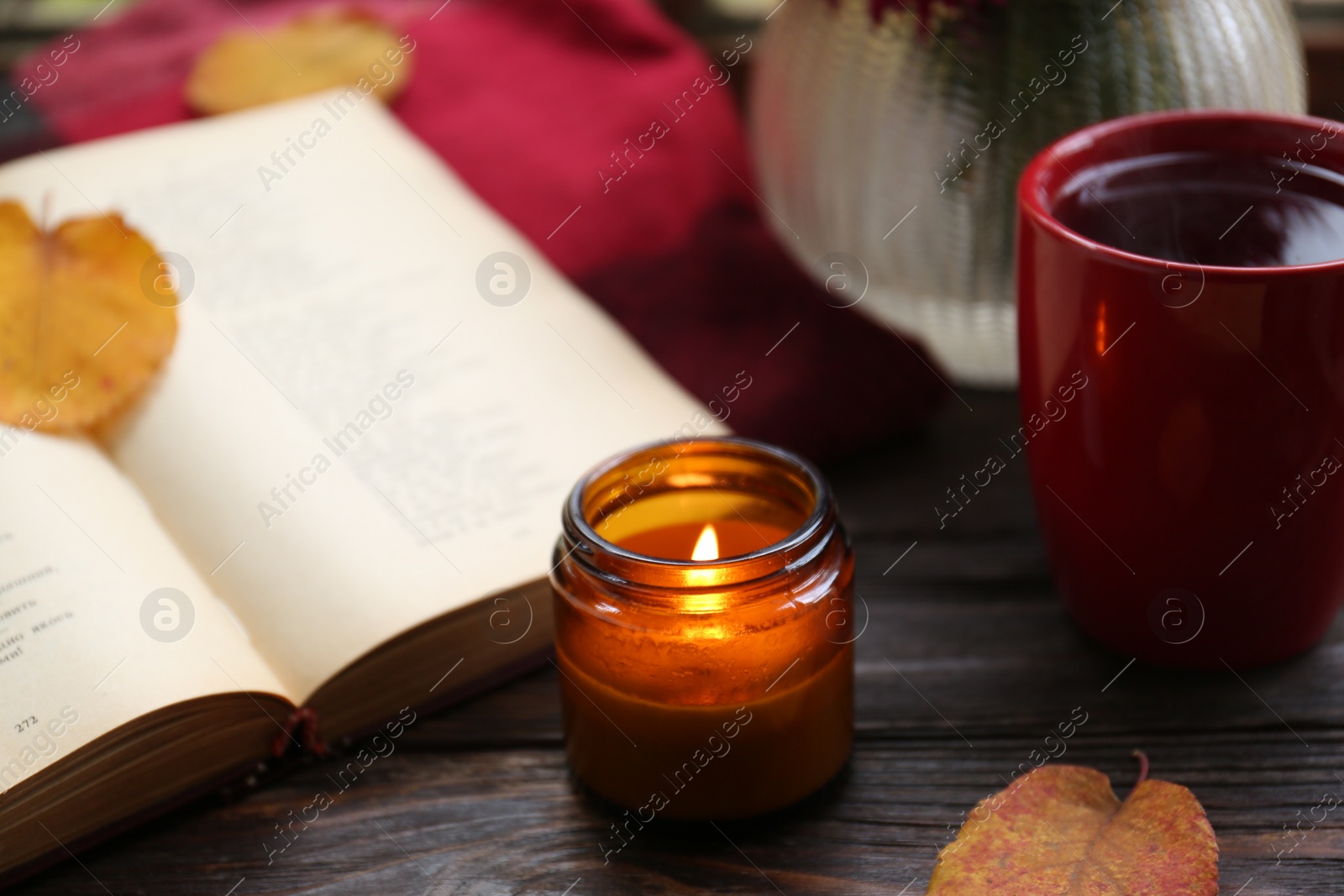 Photo of Burning scented candle, cup of tea and book on wooden table. Autumn atmosphere