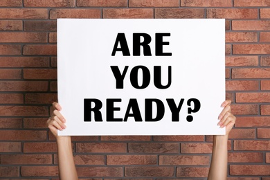 Image of Woman holding poster with text Are You Ready? near red brick wall, closeup