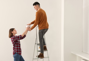 Photo of Young woman giving paint can to boyfriend on stepladder indoors. Room renovation