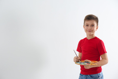 Photo of Little child with painting brush and palette near blank white wall, space for text