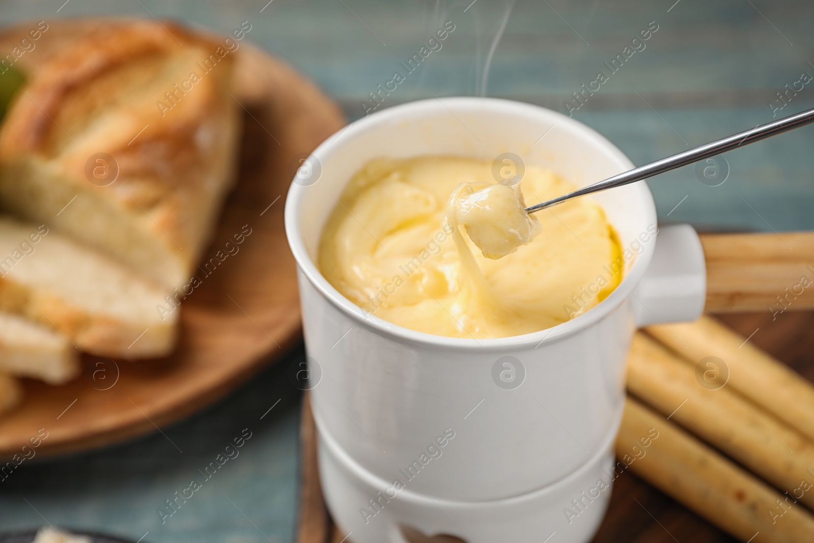 Photo of Pot of tasty cheese fondue and fork with bread on blue wooden table