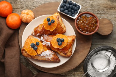 Fresh tasty puff pastry with sugar powder, jam, tangerines and blueberries on wooden table, flat lay