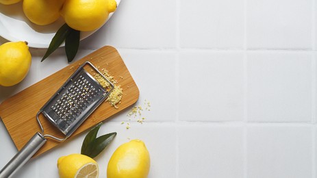 Wooden board with grater and fresh lemon zest on white tiled table, flat lay. Space for text