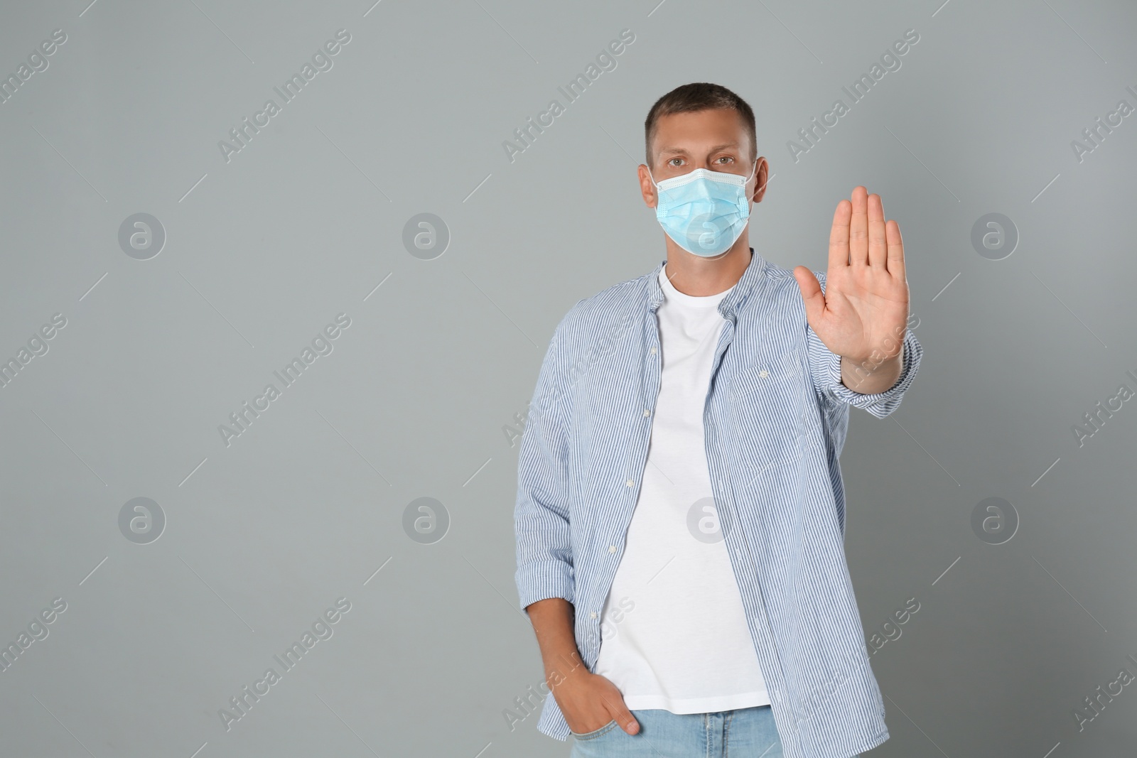 Photo of Man in protective mask showing stop gesture on grey background, space for text. Prevent spreading of coronavirus