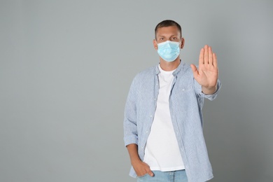 Photo of Man in protective mask showing stop gesture on grey background, space for text. Prevent spreading of coronavirus
