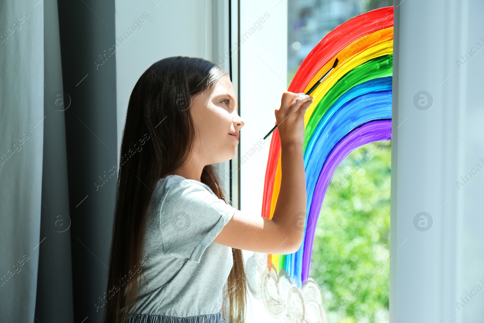 Photo of Little girl drawing rainbow on window indoors. Stay at home concept