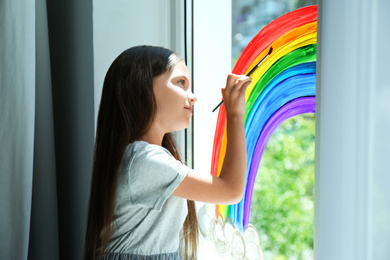 Photo of Little girl drawing rainbow on window indoors. Stay at home concept