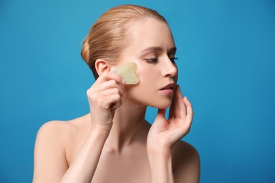 Photo of Beautiful young woman doing facial massage with gua sha tool on blue background