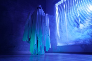 Photo of Creepy ghost. Woman covered with sheet near window in blue light, low angle view