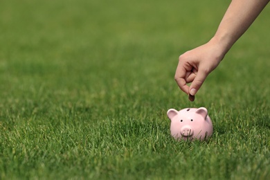 Photo of Young woman putting coin into piggy bank on green grass outdoors, closeup. Space for text