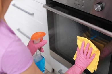 Photo of Woman cleaning oven with rag and detergent in kitchen, closeup