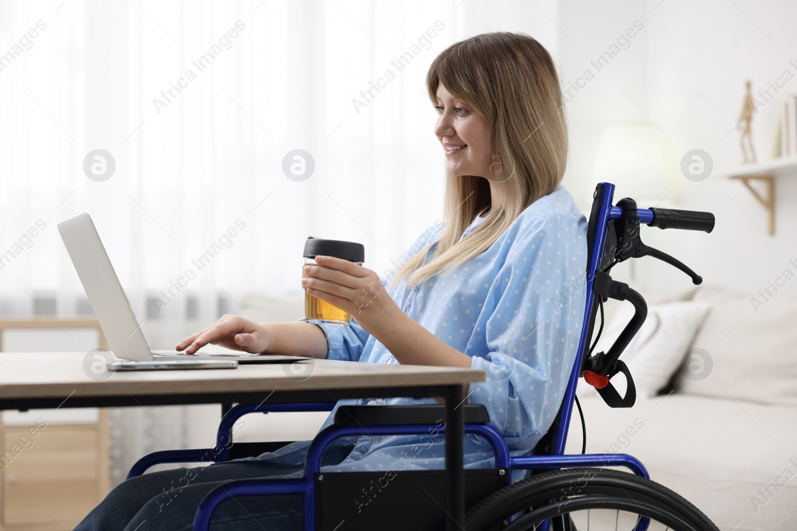 Photo of Woman in wheelchair with cup of drink using laptop at table in home office