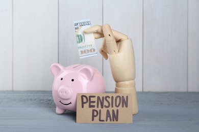 Photo of Card with phrase Pension Plan, mannequin hand holding dollar banknote and piggy bank on light blue wooden table. Retirement concept