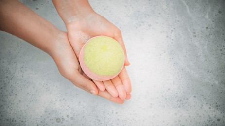 Photo of Woman holding bath bomb over water with foam, top view. Space for text