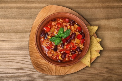 Photo of Bowl with tasty chili con carne and nachos on wooden background, top view