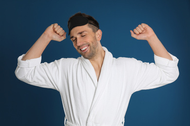 Happy young man in bathrobe and eye sleeping mask stretching on blue background