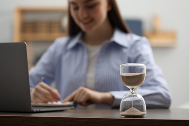 Photo of Hourglass with flowing sand on desk. Woman taking notes while using laptop indoors, selective focus