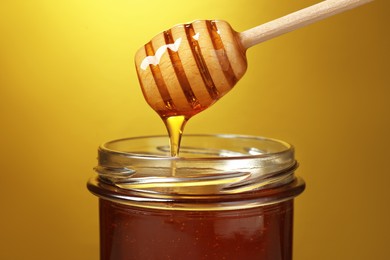Photo of Pouring tasty honey from dipper into glass jar on golden background, closeup