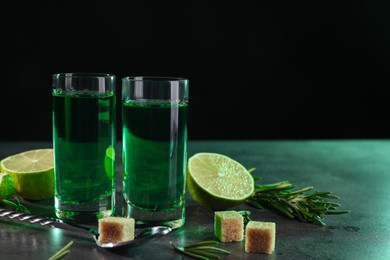 Photo of Absinthe in shot glasses, spoon, brown sugar, lime and rosemary on gray textured table, space for text. Alcoholic drink