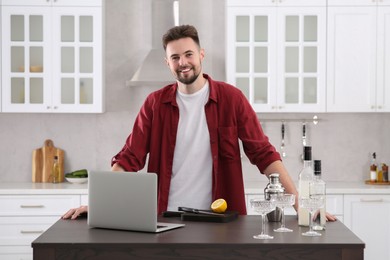 Photo of Portrait of man with cocktail ingredients and laptop at table in kitchen. Time for hobby
