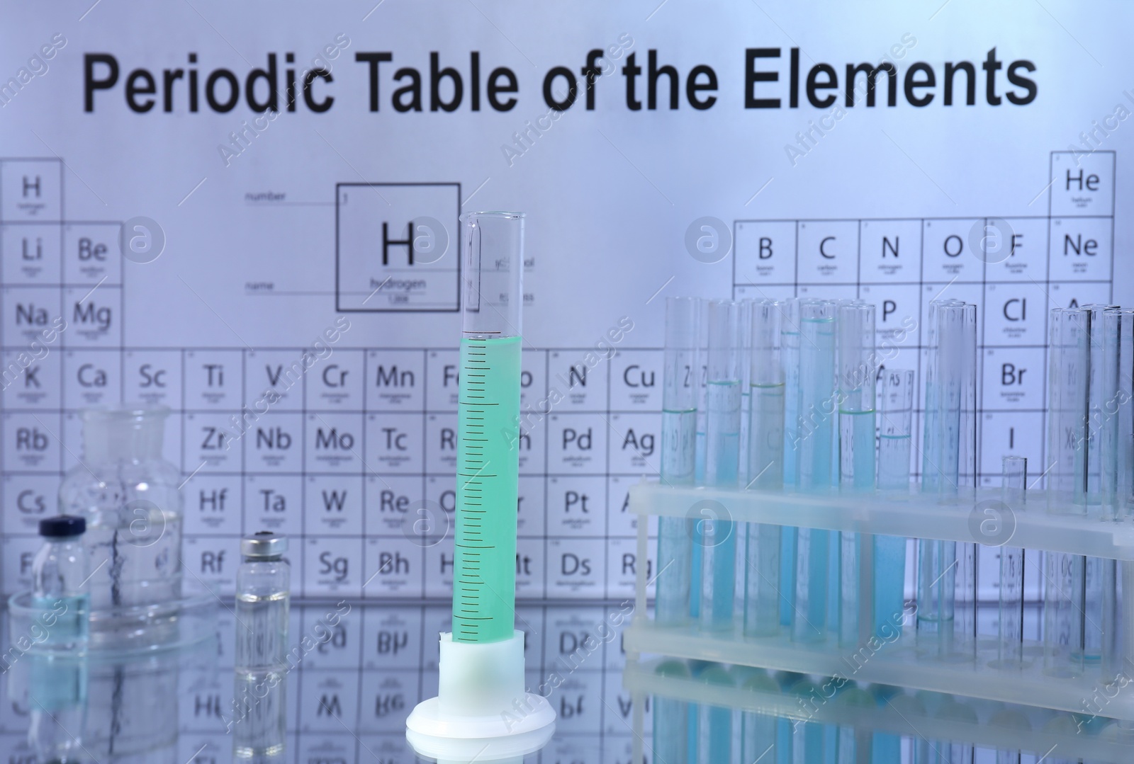 Photo of Graduated cylinder, bottles and test tubes in rack on mirror surface against periodic table of chemical elements
