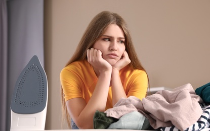 Photo of Emotional woman near board with iron and pile of clothes at home