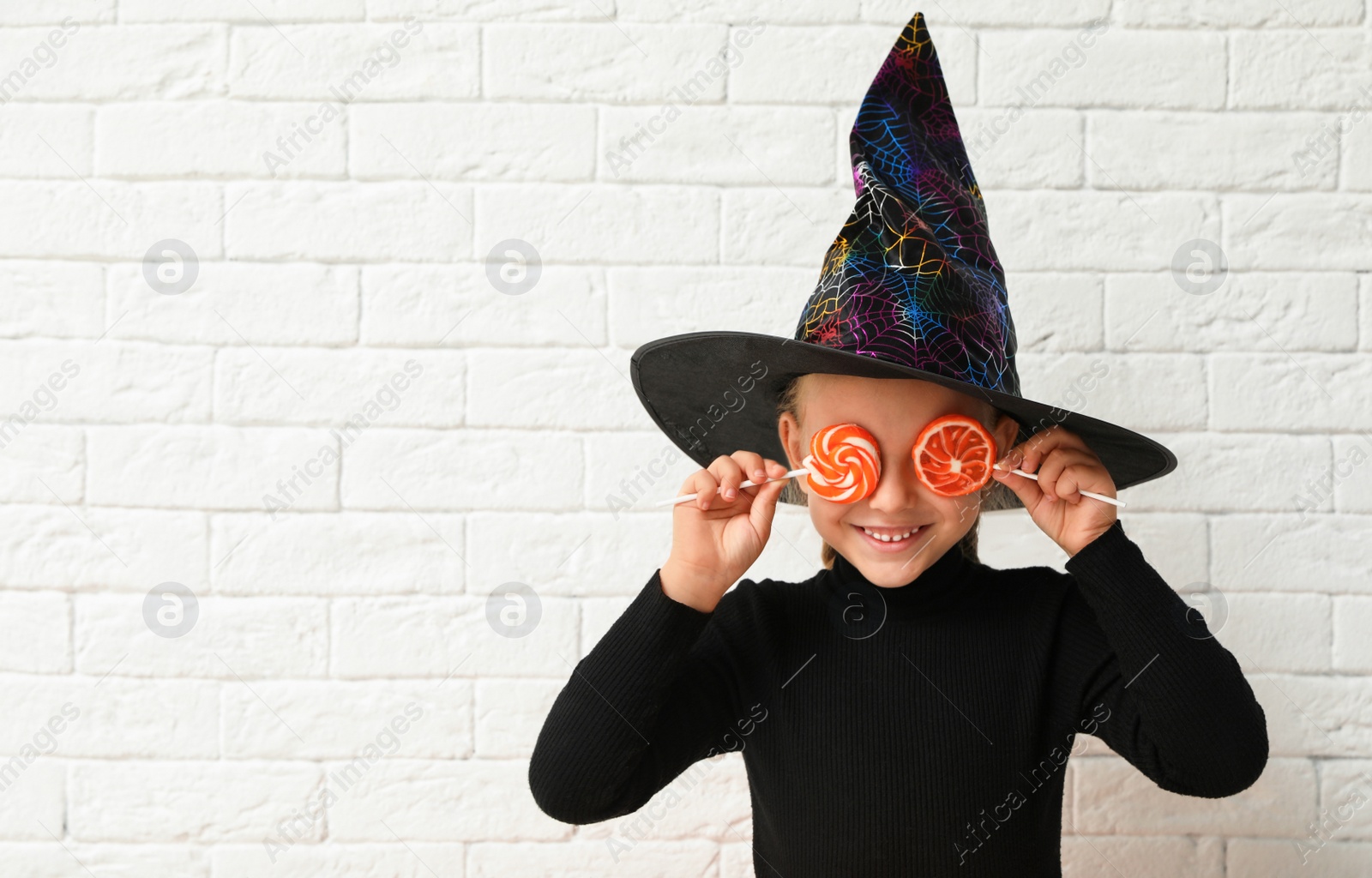 Photo of Cute little girl with candies wearing Halloween costume near white brick wall. Space for text