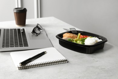 Container with tasty food, laptop and notebook on light grey table. Business lunch