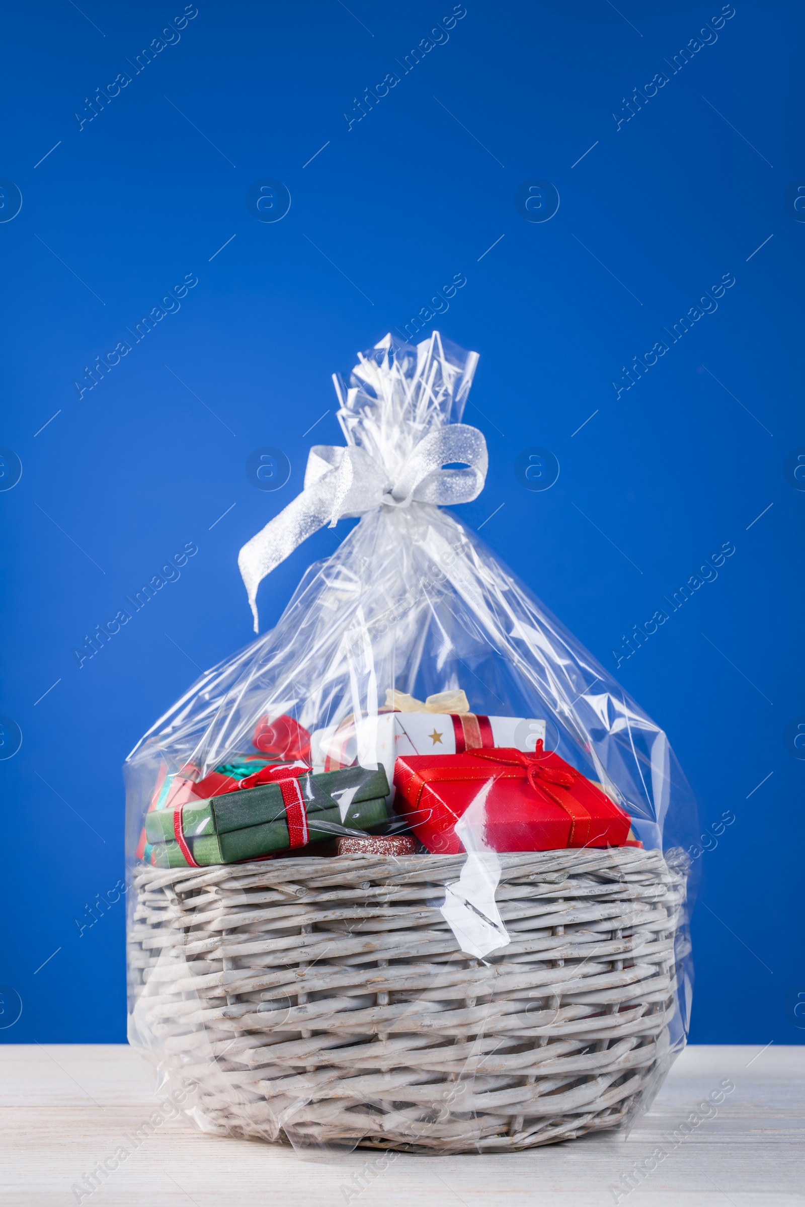 Photo of Wicker basket full of gift boxes on white wooden table against blue background