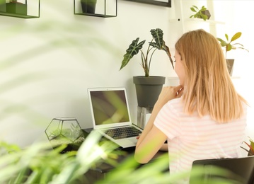 Young woman using laptop at home, space for text. Trendy room interior with plants