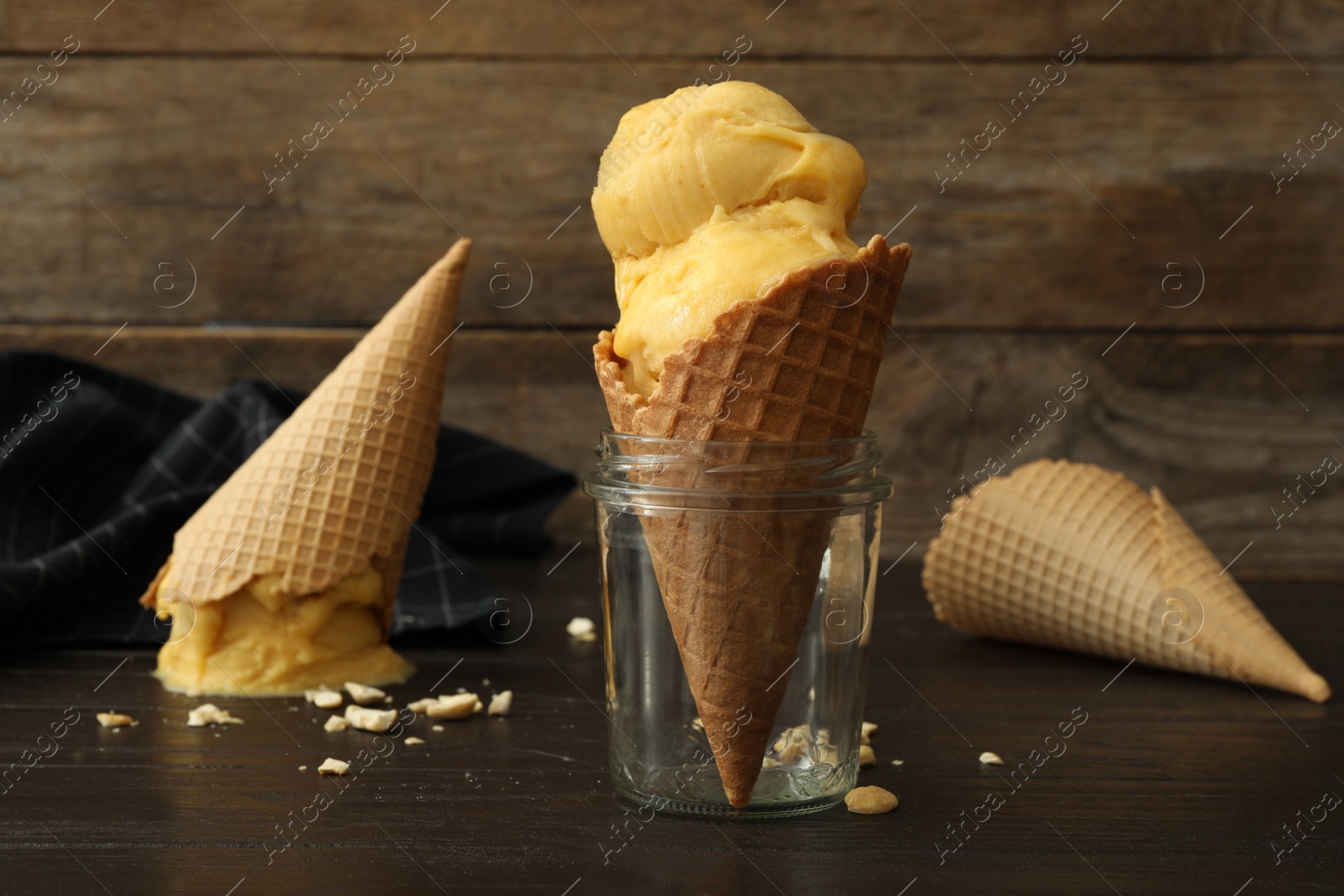 Photo of Delicious yellow ice cream in wafer cone and glass jar on black wooden table, closeup