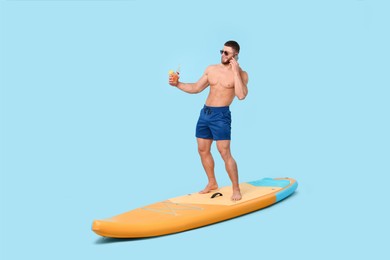 Photo of Happy man with refreshing drink talking on smartphone on SUP board against light blue background