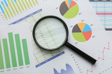 Photo of Magnifying glass on accounting documents with data and graphs, top view