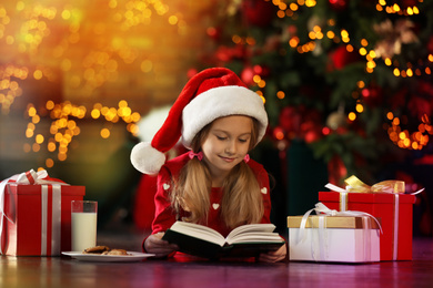 Photo of Little girl in Santa Claus cap reading book near Christmas tree indoors